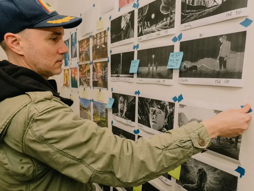 A man wearing a cap stands in front of a wall of animated stills - pictures stuck to the wall. The storyboard of a film. He is pressing the corner piece of tape on one piece of paper.
