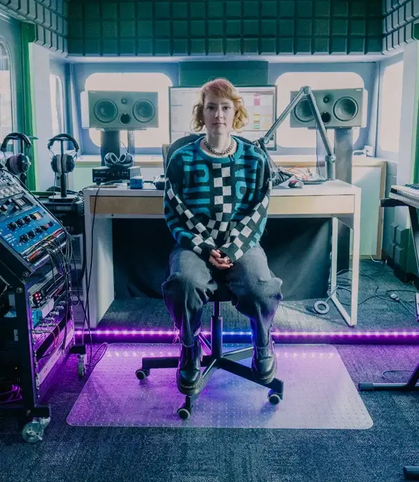 Girl sits on rolling chair in a recording studio space - lit with purple LEDs on the floor. SAE MUSIC ALUMNI, BECKI WHITTON