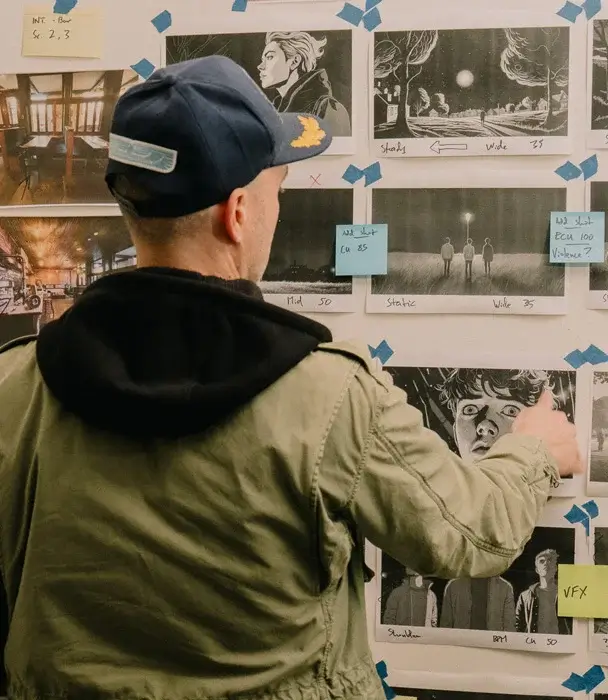 Man wearing cap and hoodie, back to camera, stands in front of wall covered with movie illustrations - storyboard/script. SAE MASTER OF CREATIVE INDUSTRIES POSTGRAD, MARK GAMBINO