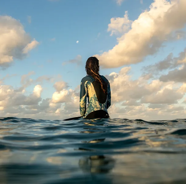 Female surfer wears a weatsuit and sits on a surfboard and in the ocean. Looking out to sea.
