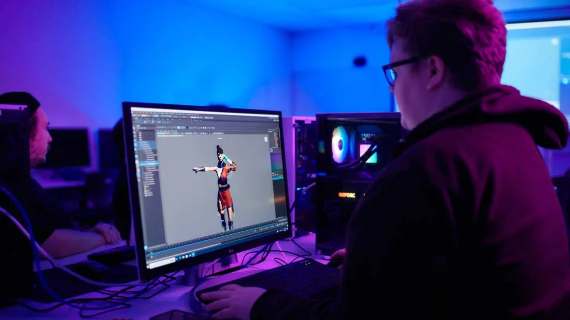 A man sits at desk and looks at a monitor displaying 3D model and editing software. The man is in a classroom. Another student sits in background and is looking at opposite wall with projected image.