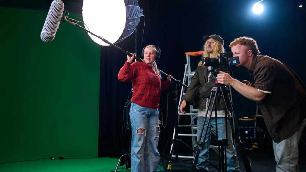 A cameraman is hunched over camera on a tripod. A man looks on in background. A girl is wearing headphones and holding a foley boom microphone over her shoulder. As soft box light is on a tripod in the background and the wall is green.