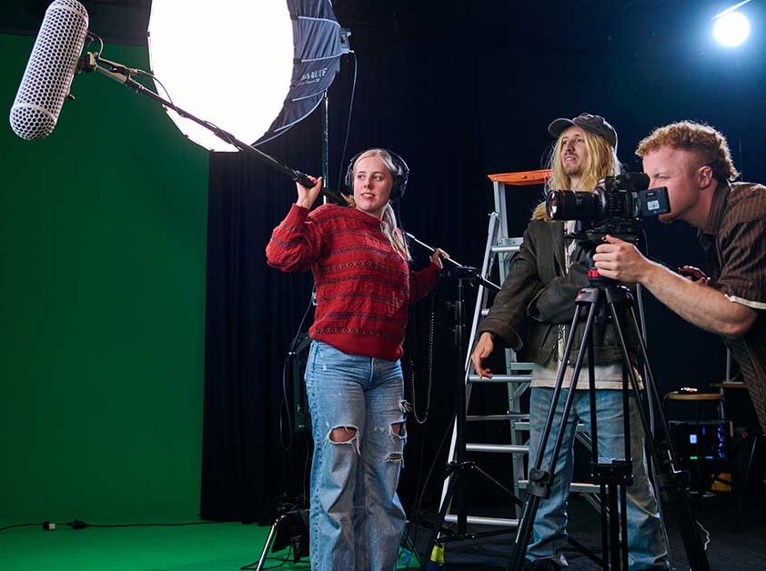 A cameraman is hunched over camera on a tripod. A man looks on in background. A girl is wearing headphones and holding a foley boom microphone over her shoulder. As soft box light is on a tripod in the background and the wall is green.