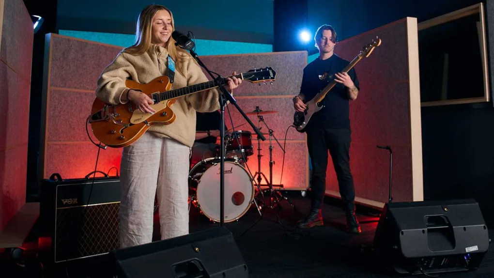A girl plays guitar in front of microphone and man in background on bass guitar. Both people are in a recording studio with amplifier and drums in the background.