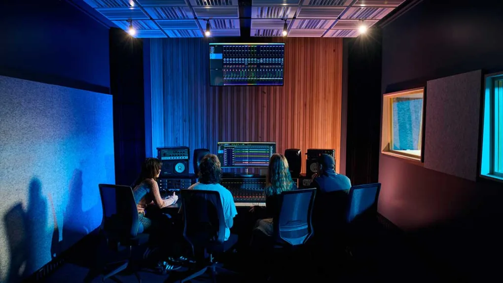 A group of people sit in chairs looking at a wall with a screen on it. They face away from camera towards a mixing console audio desk.