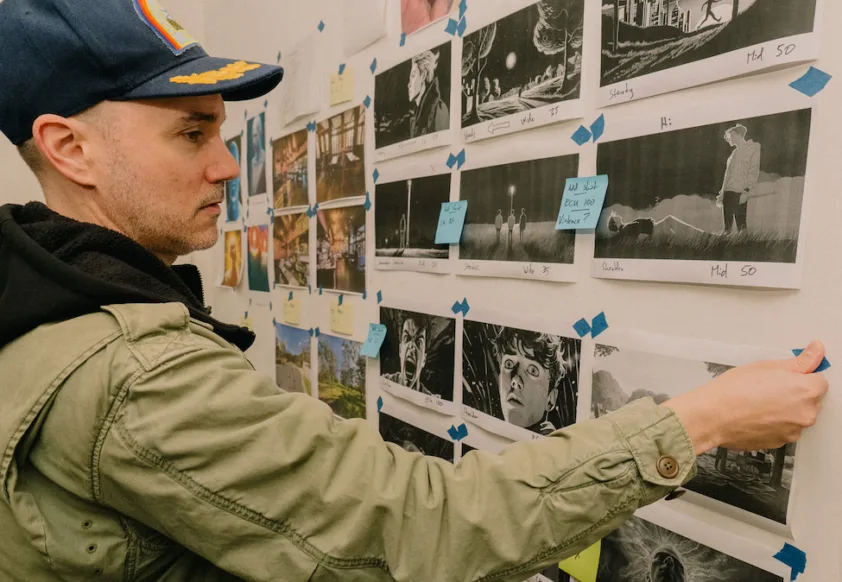 Man wearing cap and hoodie, back to camera, stands in front of wall covered with movie illustrations - storyboard/script. SAE MASTER OF CREATIVE INDUSTRIES POSTGRAD, MARK GAMBINO