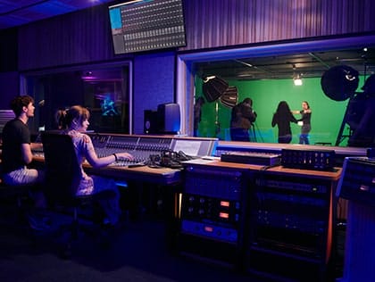 Two students sitting in a production booth, with performers in the background tile thumbnail