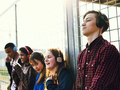 A young man wearing over-ear headphones, with four other students in the background tile thumbnail