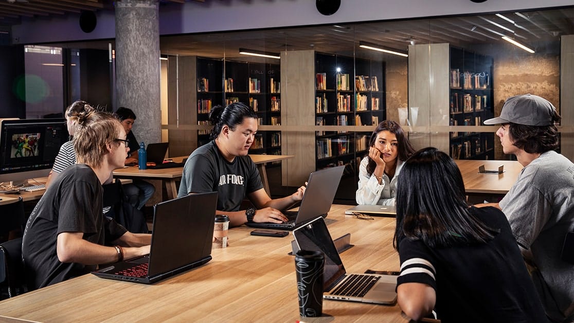 Five students sitting around a desk facing each other, with laptops open desktop