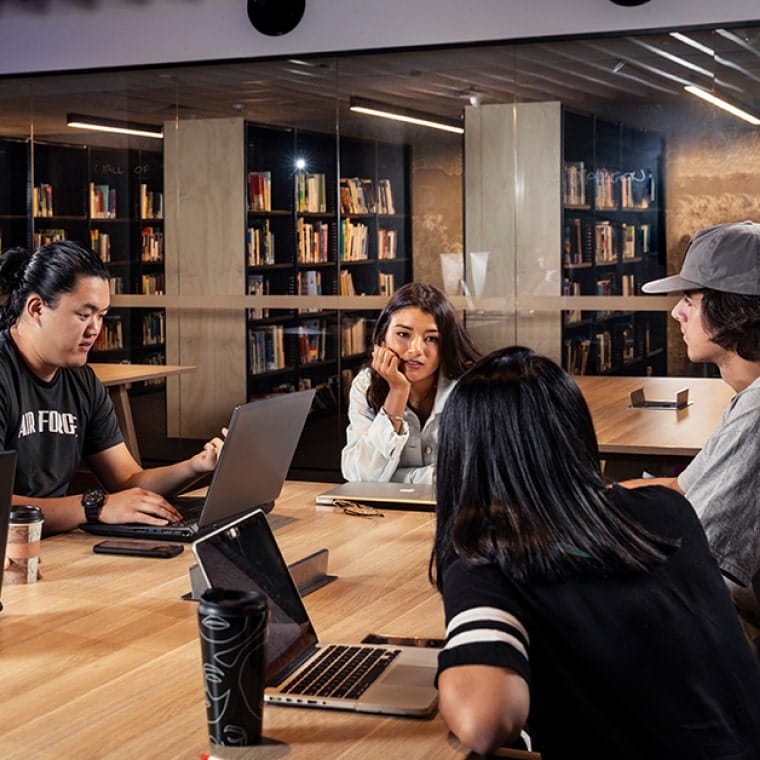 Five students sitting around a desk facing each other, with laptops open desktop