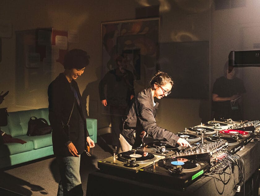 Two people stand over a mixing desk - filled with nine turntables and a mixing console. People are seen in the shadows in the background.