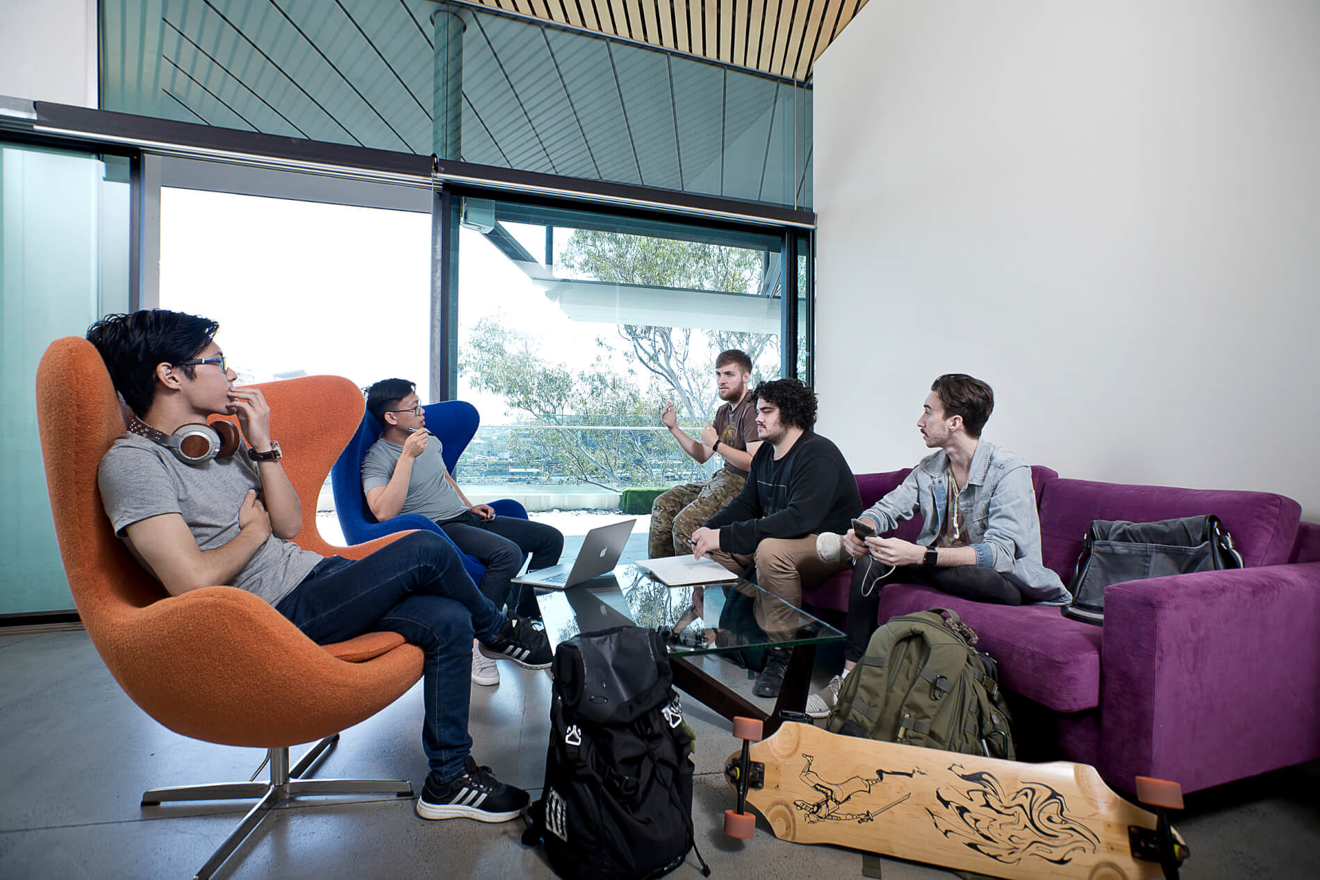 A group of students sit around coffee table. Some in chairs. Some on a couch. All in conversation.