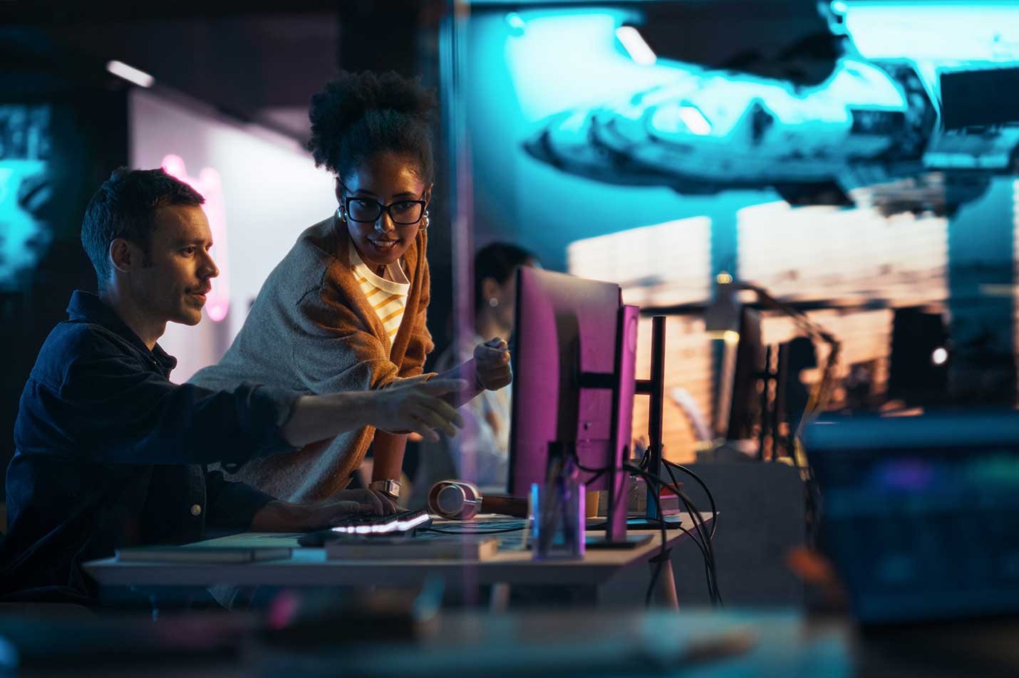 A man sitting and woman standing at a desk in a backlit room, pointing towards a computer monitor