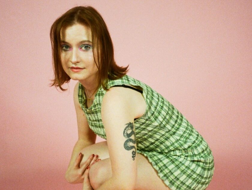 Photo of Amelia Thompson, leaning down with her hands wrapped around her knees in front of a pink backdrop