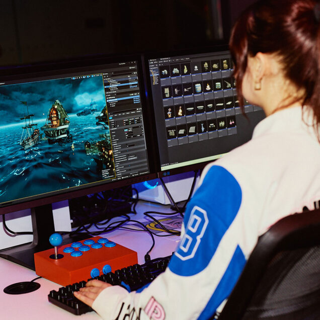 A woman with red hair in a ponytail facing away from camera, working with games software on a desktop monitor