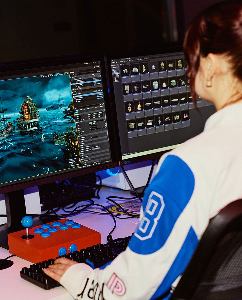 A woman with red hair in a ponytail facing away from camera, working with games software on a desktop monitor