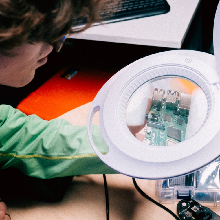 Man looks through magnifying lens. A computer motherboard with integrated circuits shown in lens. A keyboard is seen in background on a desk.