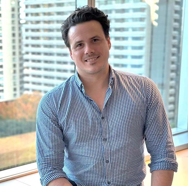 Picture of man smiling to camera. Sits in office in front of window. Office buildings in background. SAE lecturer and teacher, Dr Luke Bozzetto