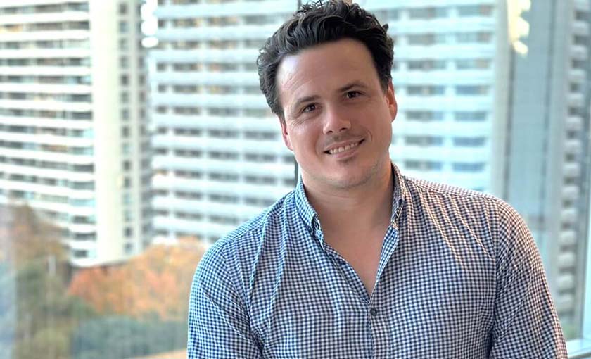 Picture of man smiling to camera. Sits in office in front of window. Office buildings in background. SAE lecturer and teacher, Dr Luke Bozzetto