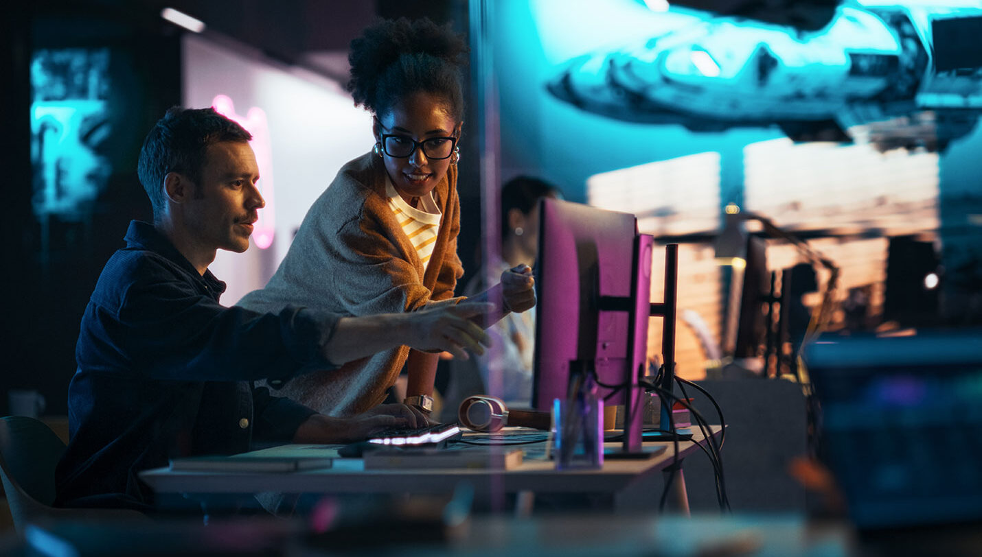 A man sitting and woman standing at a desk in a backlit room, pointing towards a computer monitor