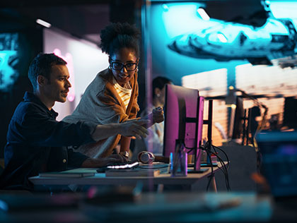 A man sitting and woman standing at a desk in a backlit room, pointing towards a computer monitor