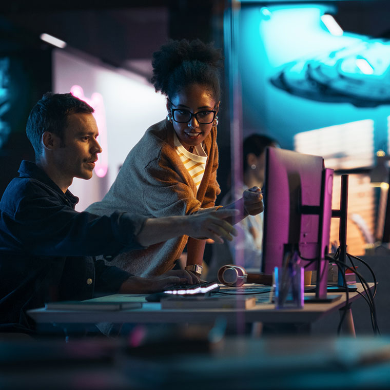 A man sitting and woman standing at a desk in a backlit room, pointing towards a computer monitor