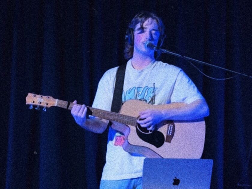 You man in jeans and T-shirt sings into a microphone while playing guitar on a dark stage