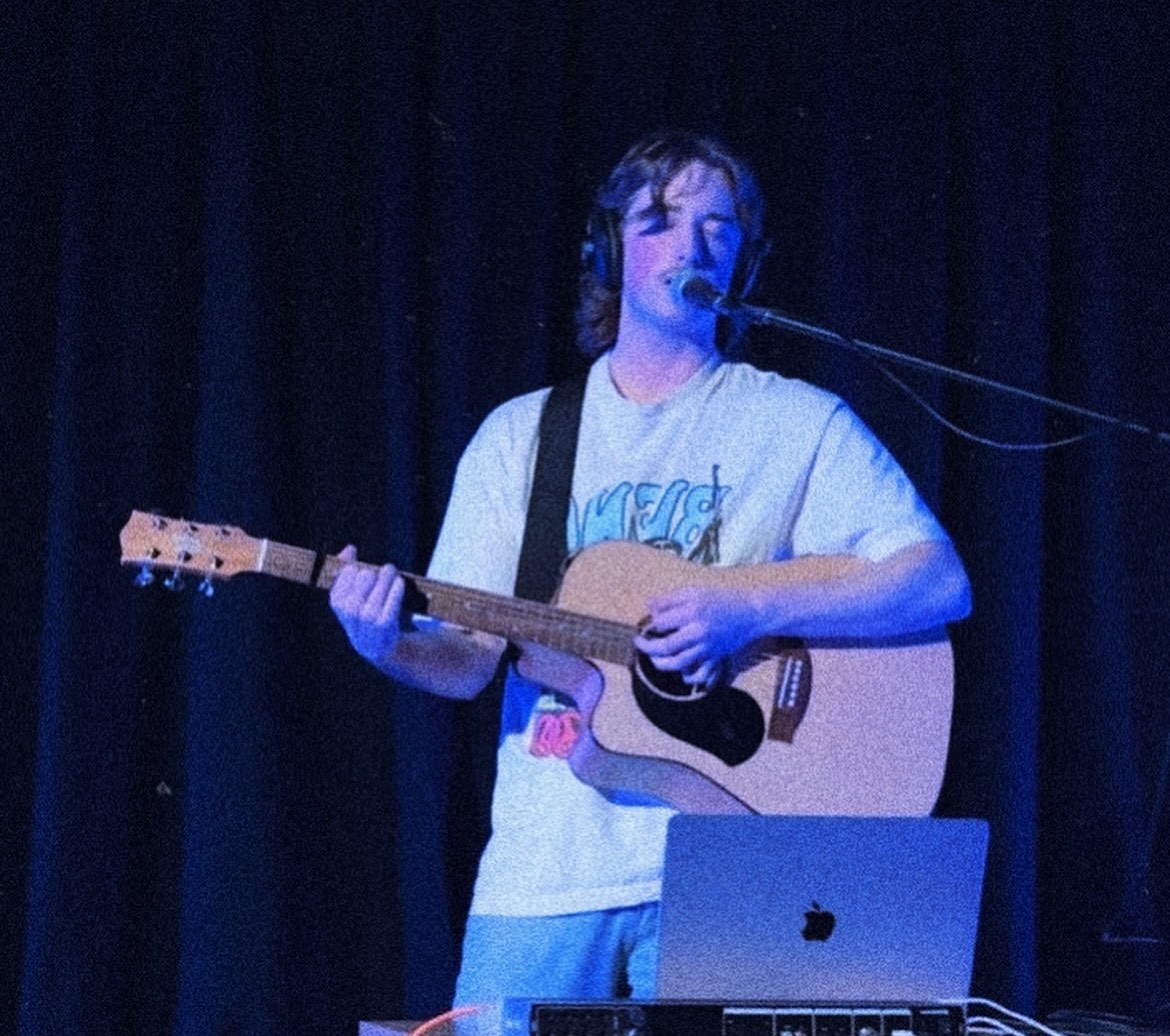 You man in jeans and T-shirt sings into a microphone while playing guitar on a dark stage