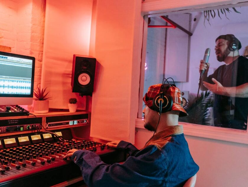 Two men in a sound studio. Background man in sound booth with headphones and at microphone. Man in foreground sitting with headphones on in front of audio mixing console. Monitor with sound editing software on screen.