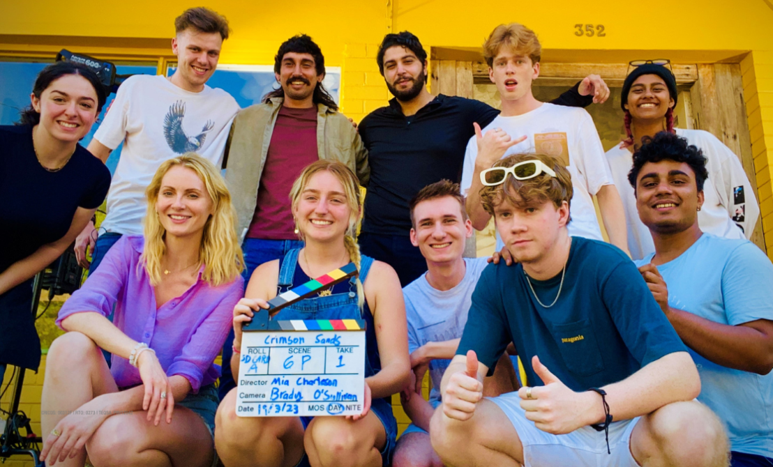 eleven young men and women smile at the camera on the porch of a house holding a film card