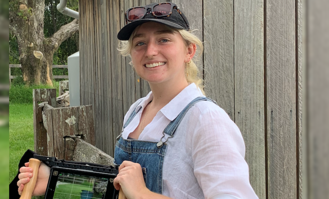 young woman with blonde hair in a cap and sunglasses on forehead wearing overalls smiles at camera holding a filming tool