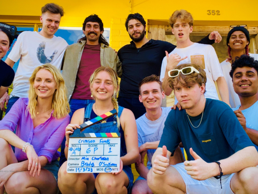 eleven young men and women smile at the camera on the porch of a house holding a film card