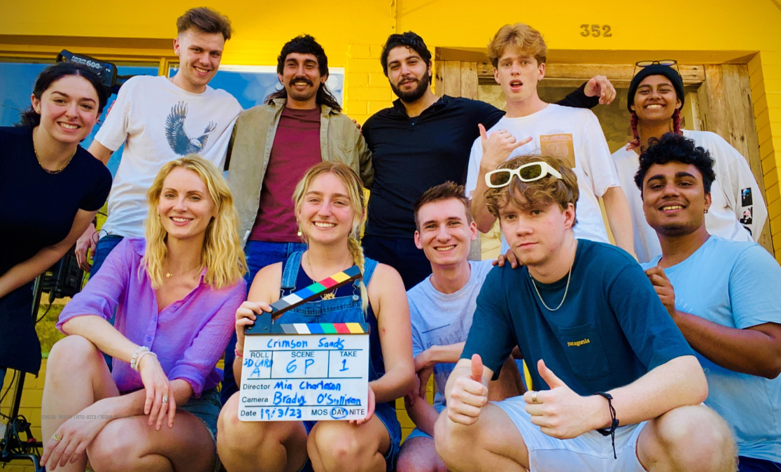 eleven young men and women smile at the camera on the porch of a house holding a film card