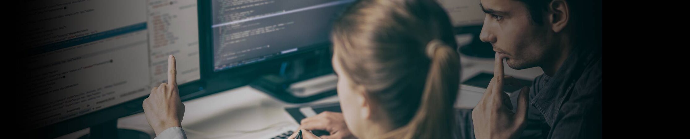 Two people sit in front of monitor and code on dual screens.