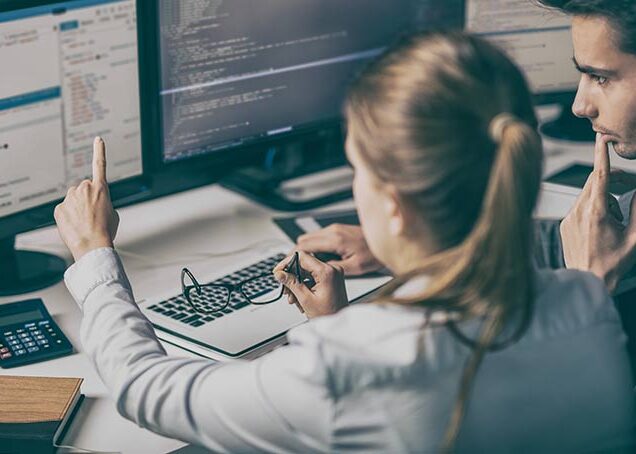 Two people sit in front of monitor and code on dual screens.