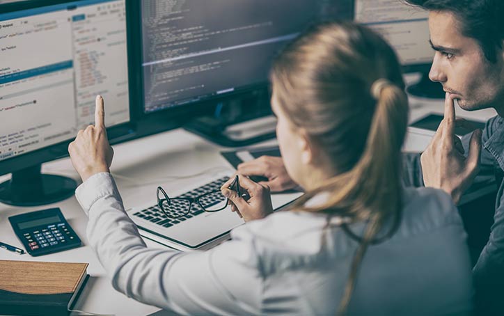 Two people sit in front of monitor and code on dual screens.