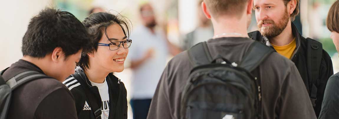 Group of four people stand speaking and facing each other. All wear backpacks. Smiling. Outdoor environment.