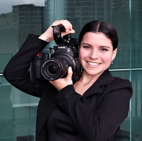 Student smiles at camera with a professional quality film camera on her shoulder