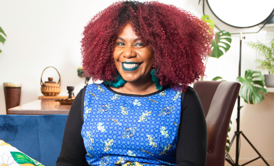 Woman with red afro hair and green lipstick and earrings gives the camera a big smile