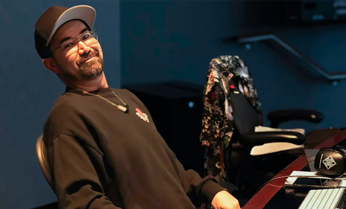 man leans back in chair in an office with cap and glasses on with a small smile