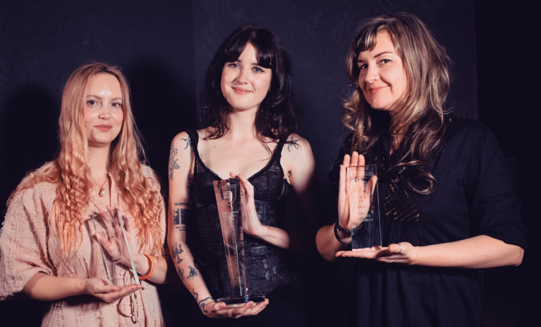 Three women stand holding trohpies in front of a black background in evening attire