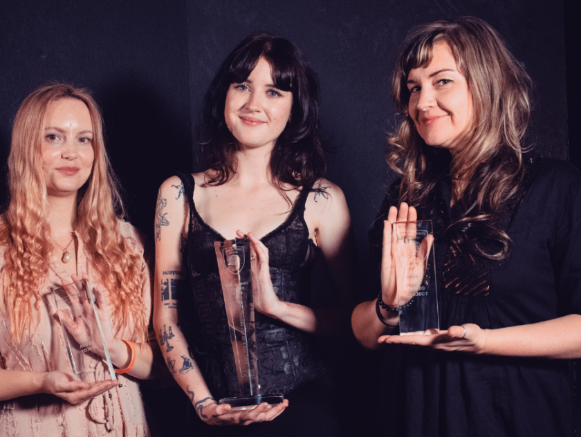 Three women stand holding trohpies in front of a black background in evening attire