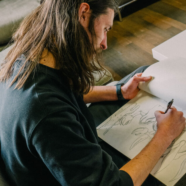 A man is holding a sketchpad and drawing on the pad with pen and looking down