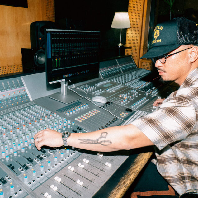 Man wearing a hat and spectacle sits with outstretched arms on audio console.