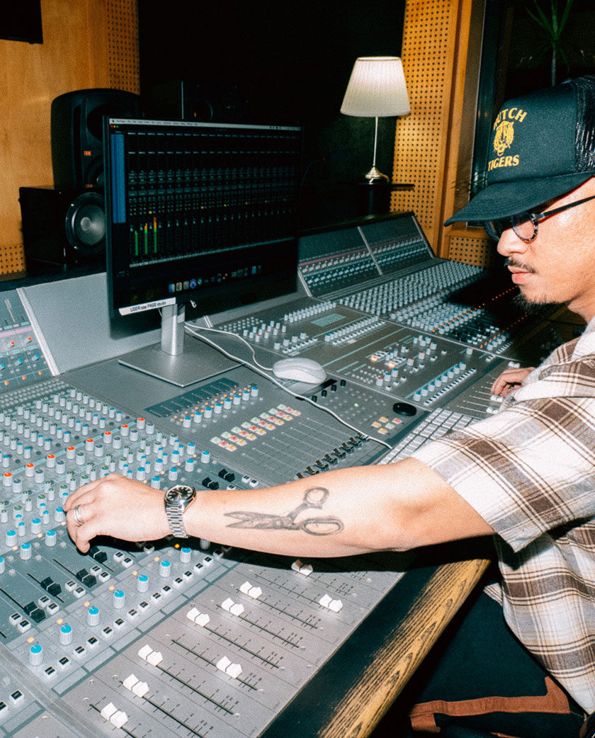 Man wearing a hat and spectacle sits with outstretched arms on audio console.