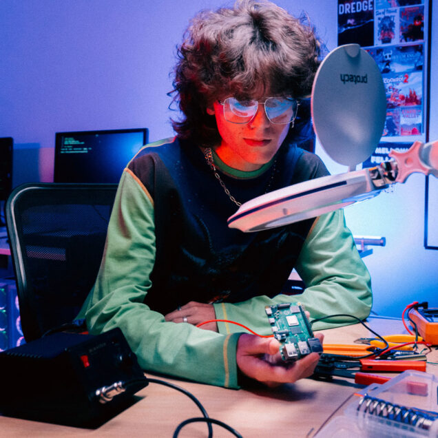 Man wearing glasses sit in front of magnifying glass holding a motherboard. A solder machines and pliers on desk