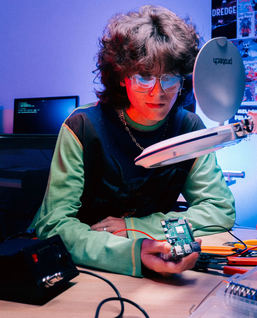 Man wearing glasses sit in front of magnifying glass holding a motherboard. A solder machines and pliers on desk