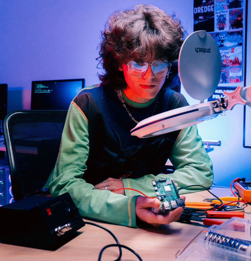 Man wearing glasses sit in front of magnifying glass holding a motherboard. A solder machines and pliers on desk
