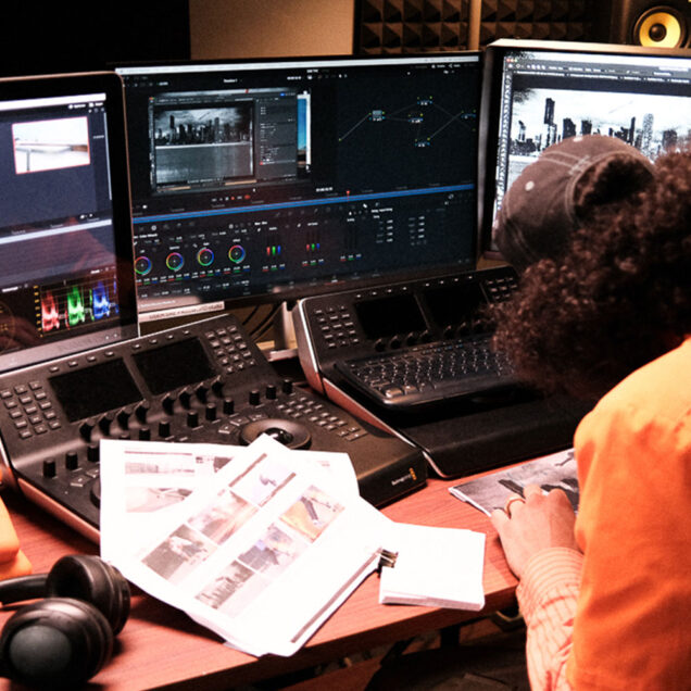 Three monitors are in front on desk. Two mixing consoles in front of person looking down at paper. Paper on table