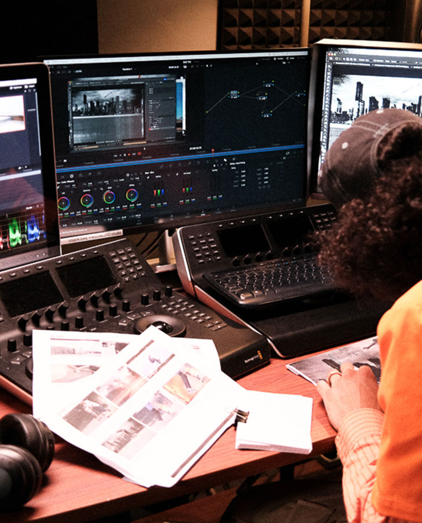 Three monitors are in front on desk. Two mixing consoles in front of person looking down at paper. Paper on table